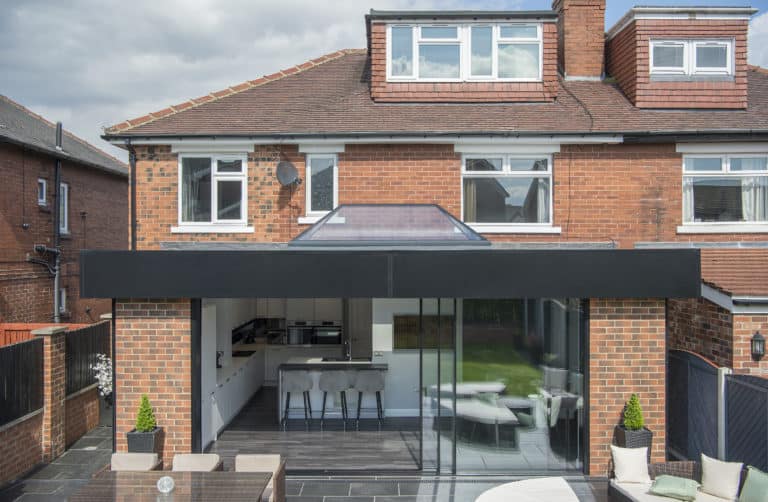 A modern brick house featuring bespoke glass solutions in the kitchen extension, sliding doors opening to a patio with tables, chairs, and potted plants. The flat roof and upper-floor dormer windows complete the contemporary look.