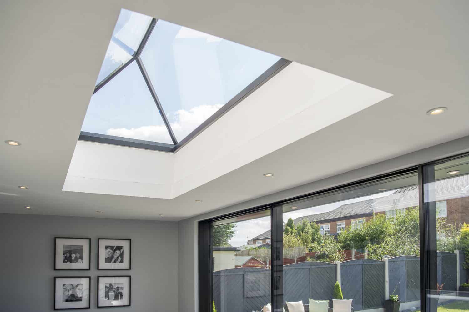 A modern living room with a large glass skylight in the ceiling, recessed lighting, bespoke glass solutions like floor-to-ceiling bifold doors, and framed photos on the wall, offering a view of a fenced backyard and neighboring houses.