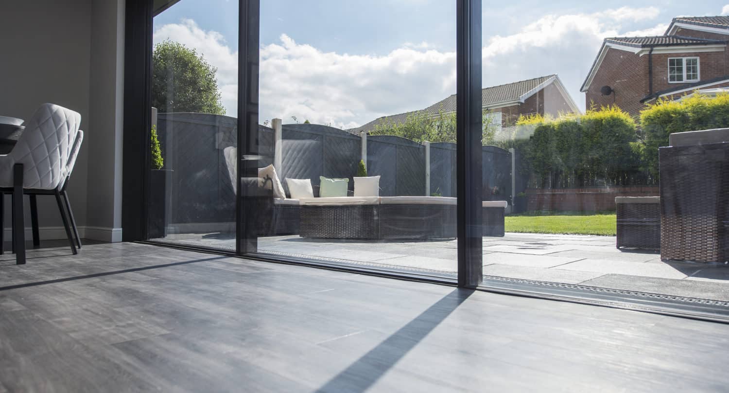View from inside a modern home through expansive bifold doors, looking out onto a sunny patio with outdoor seating, greenery, and neighboring houses in the background.