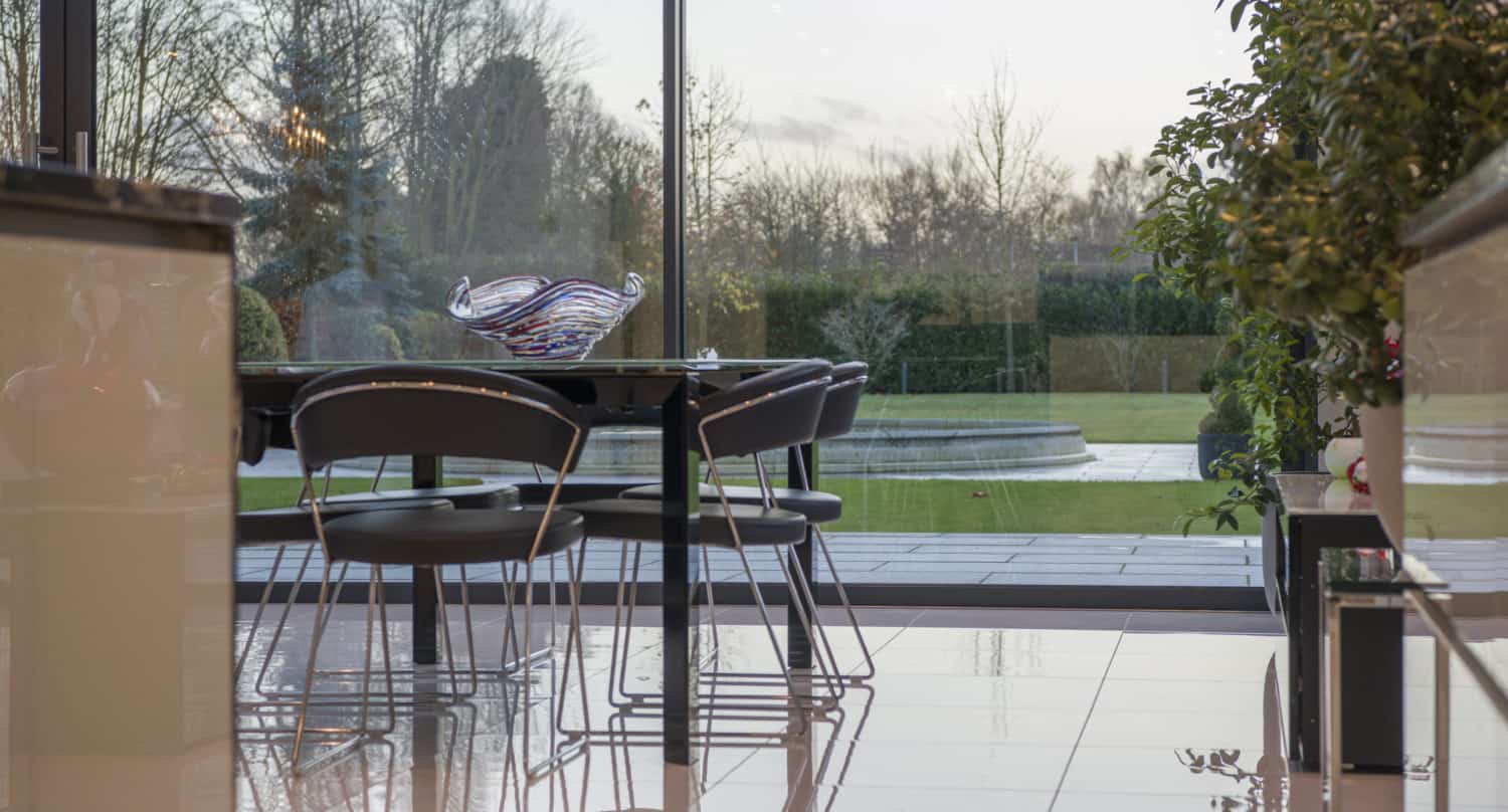 Modern dining area with four black chairs around a table, glass bowl centerpiece, and shiny white tiled floor. Large bifold doors open to a landscaped garden and patio, seamlessly blending indoor comfort with outdoor beauty.