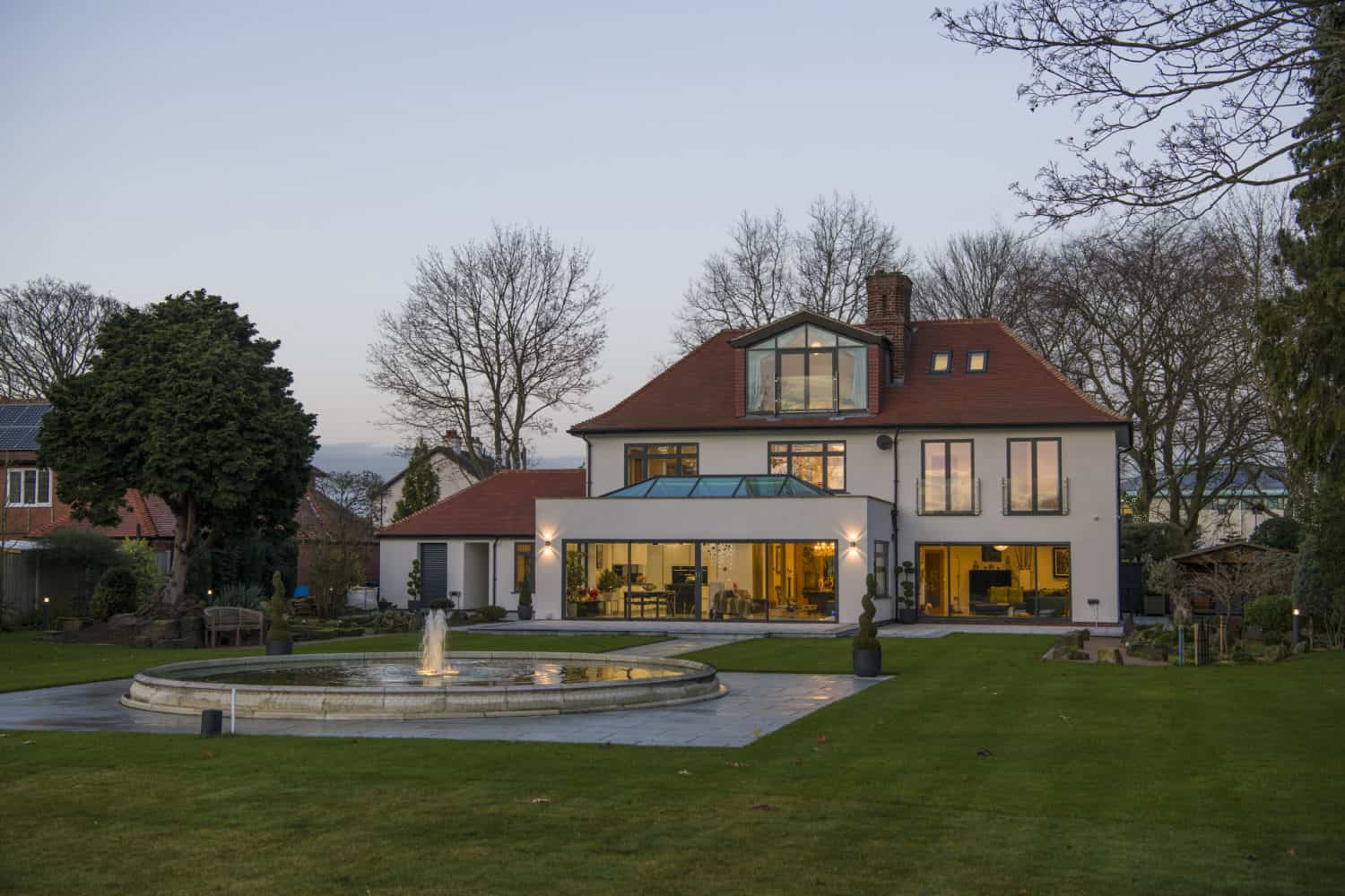 A modern two-story house with large windows, a red roof, and a glass extension featuring sleek bifold doors sits behind a circular fountain on a well-kept lawn, surrounded by trees and garden features at dusk.