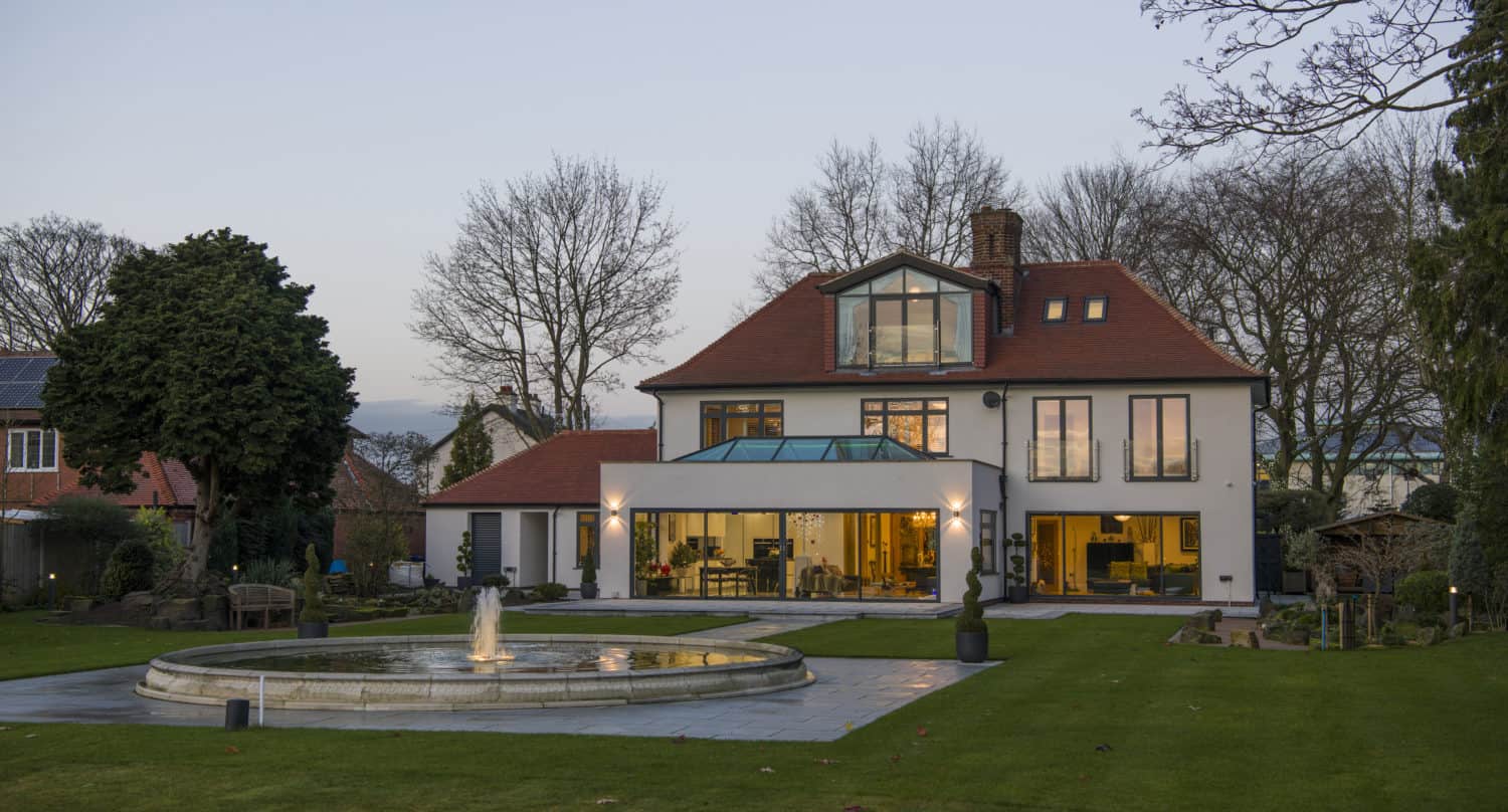 A modern two-story house with large windows, a red roof, and a glass extension featuring sleek bifold doors sits behind a circular fountain on a well-kept lawn, surrounded by trees and garden features at dusk.