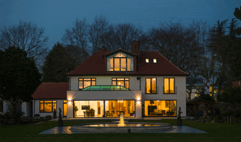 A large, modern house with many windows and sliding doors brightly lit from within at dusk, featuring a red roof and a circular fountain with water jets in the front yard. Trees without leaves stand behind the house.