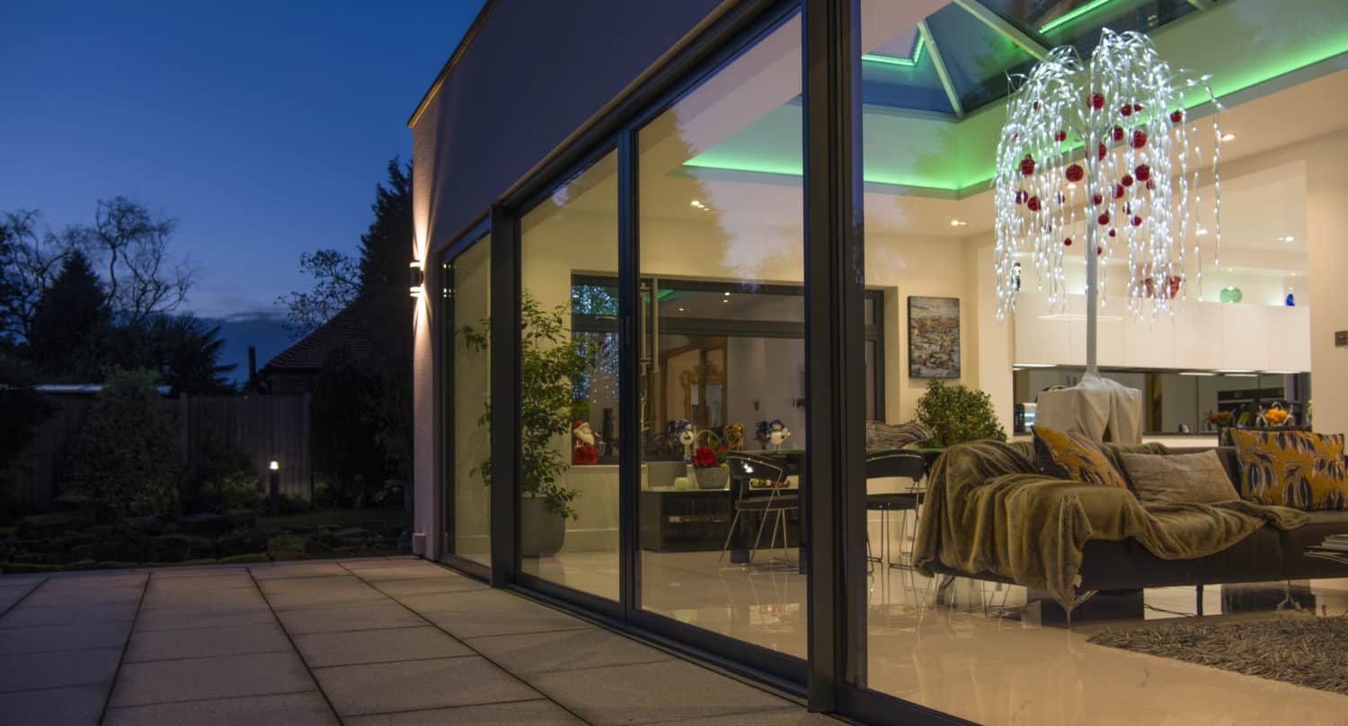 Modern house with large bifold doors revealing a stylish, well-lit living room at night; green accent lighting and a decorative chandelier are visible inside, with a spacious patio outside.