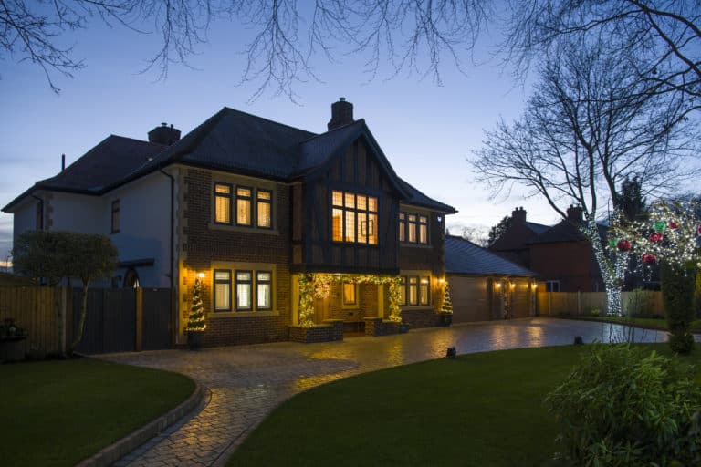 Two-story brick house with bespoke glass solutions like bifold doors and glass roofing, exterior lights glowing at dusk, a large driveway, and decorated trees with string lights and red ornaments. The sky is clear and the garden is neatly landscaped.