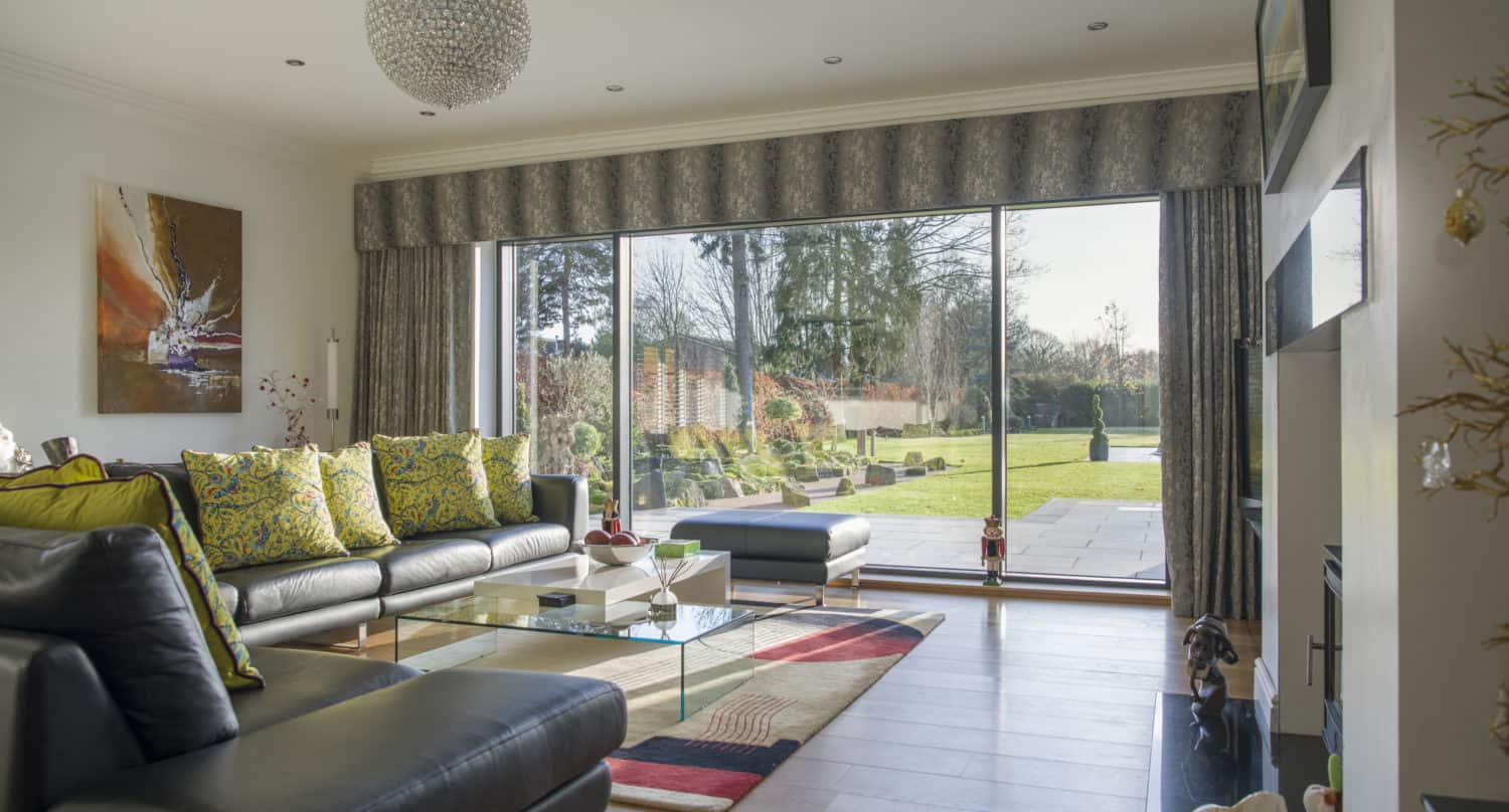 Modern living room with bespoke glass solutions, including large sliding doors opening to a sunny garden. Black leather sofas with yellow patterned pillows, glass coffee table, and colorful rug on wooden floor. Bright and airy atmosphere.