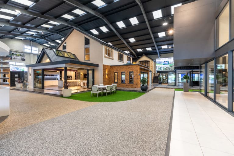 A spacious indoor showroom featuring model homes with modern architecture, bifold doors, outdoor dining furniture, large windows, and bright overhead lighting, creating a realistic residential display.