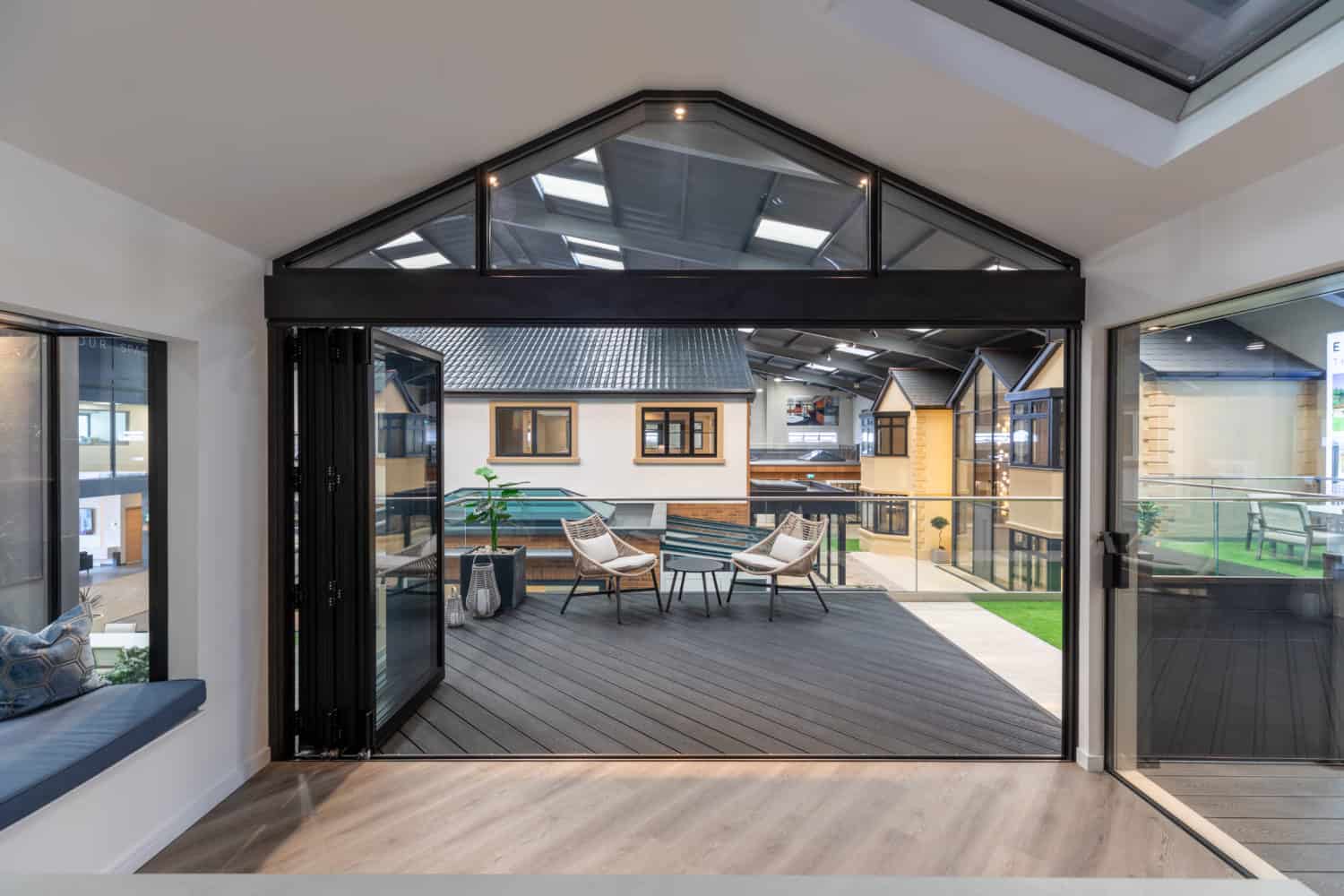 Modern indoor space with large sliding glass doors opening to a balcony with outdoor seating, overlooking a courtyard featuring contemporary buildings and striking glass roofing.