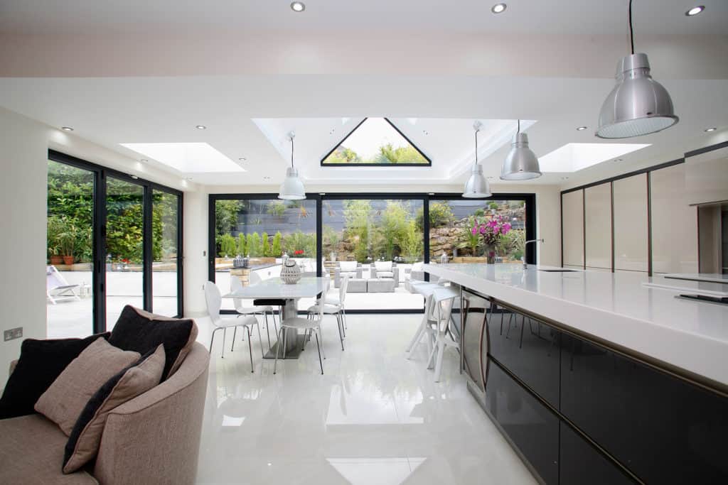 Modern open-plan kitchen and dining area with white glossy floors, large skylights, hanging pendant lights, black-framed bifold doors, a white table with chairs, and a view of a lush garden outside.