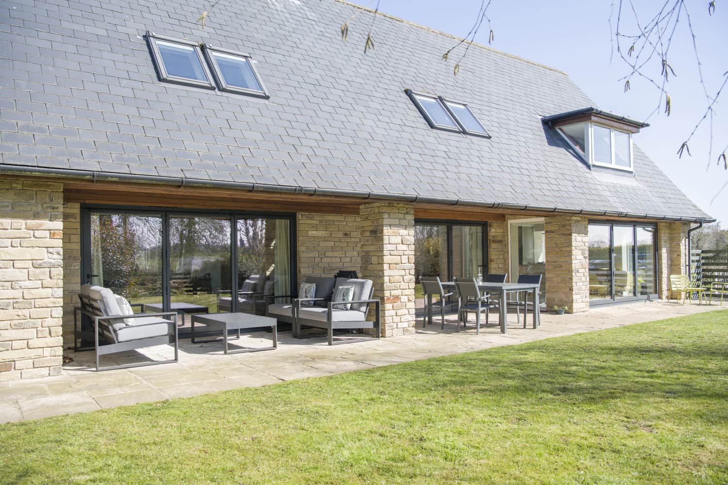 Modern house with a slate roof, large windows, and stone walls. Sliding doors open onto a patio with outdoor seating, tables, and chairs, overlooking a green lawn on a sunny day.