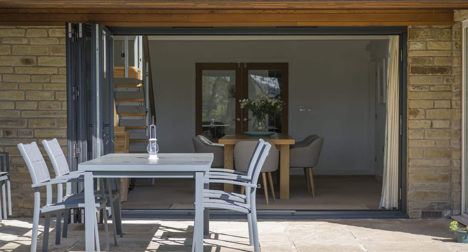 A patio with a gray table and four chairs sits outside large bifold doors, which open into a modern dining area with a wooden table, chairs, and a staircase. The exterior wall is made of stone.