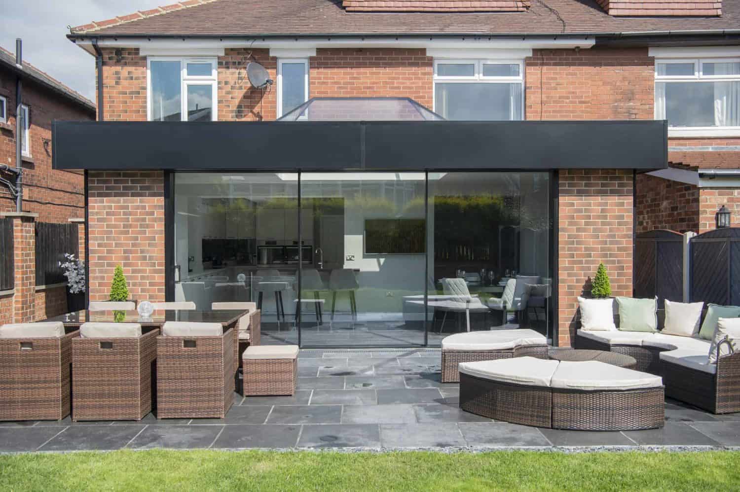 Modern brick house with large sliding doors opening to a patio with wicker outdoor furniture, grey tiles, and a green lawn in the foreground. Bespoke glass solutions offer an inviting view of the interior kitchen through the glass.