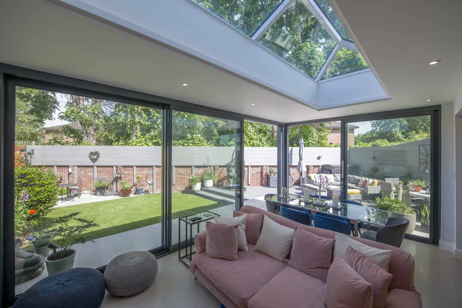 A modern living room with a pink sofa and large sliding doors opens to a sunny, landscaped backyard with patio seating, potted plants, and bespoke glass solutions like a skylight overhead that fills the space with natural light.