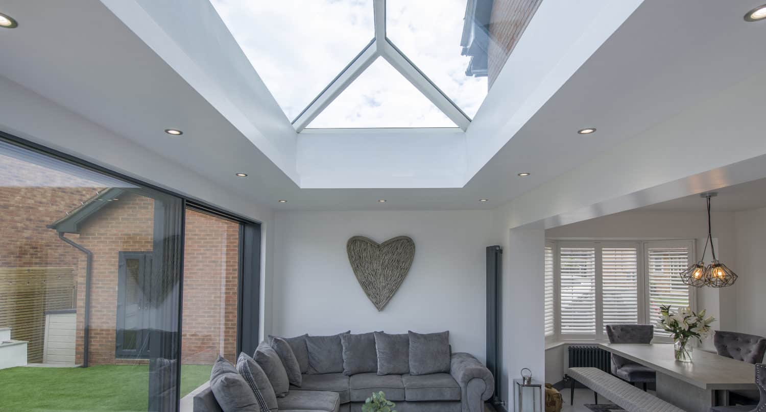 A modern living room with a large glass skylight, gray sectional sofa, heart-shaped wall decor, and glass coffee table features sliding doors that lead to an adjacent dining area with a bench and flower arrangement. White walls and recessed lighting enhance the space.
