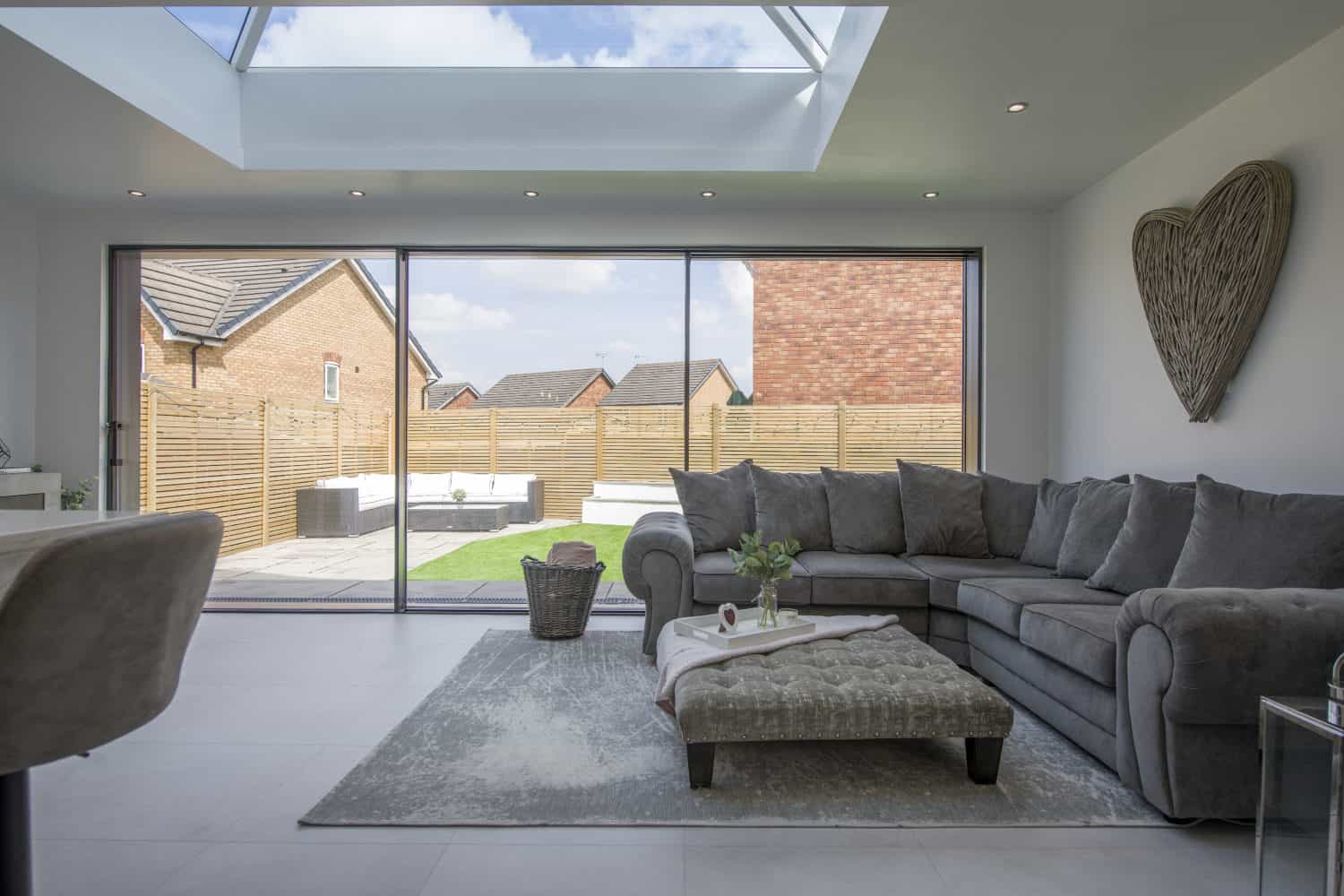 Modern living room with large gray sectional sofa, ottoman, and rug. Bespoke glass sliding doors open to a fenced backyard. Skylight above lets in natural light. Wicker heart decor hangs on the white wall.