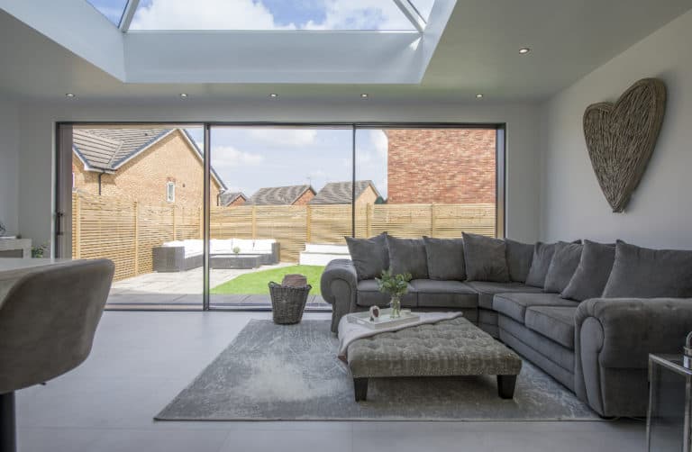 Modern living room with large gray sectional sofa, ottoman, and rug. Bespoke glass sliding doors open to a fenced backyard. Skylight above lets in natural light. Wicker heart decor hangs on the white wall.