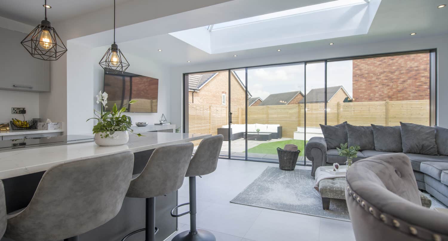 Modern open-plan kitchen and living room with grey bar stools, pendant lights, a glass roofing skylight, and sliding glass doors opening to a backyard with lawn and fencing. A grey sofa and coffee table are in the living area.