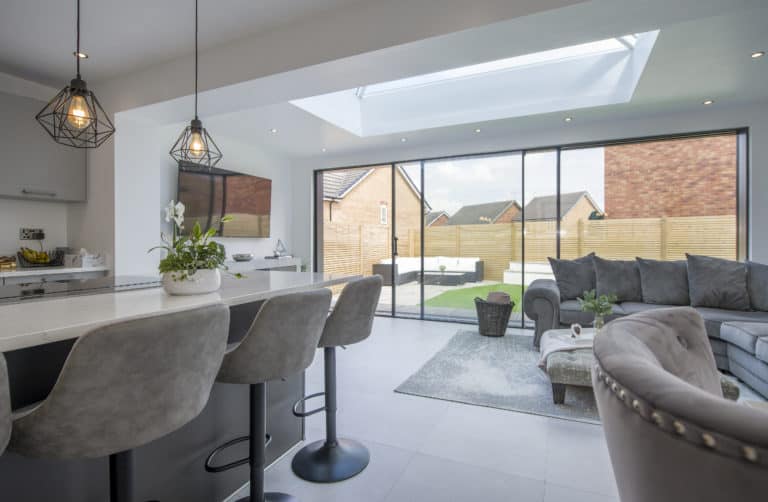 Modern open-plan kitchen and living room with grey bar stools, pendant lights, a glass roofing skylight, and sliding glass doors opening to a backyard with lawn and fencing. A grey sofa and coffee table are in the living area.