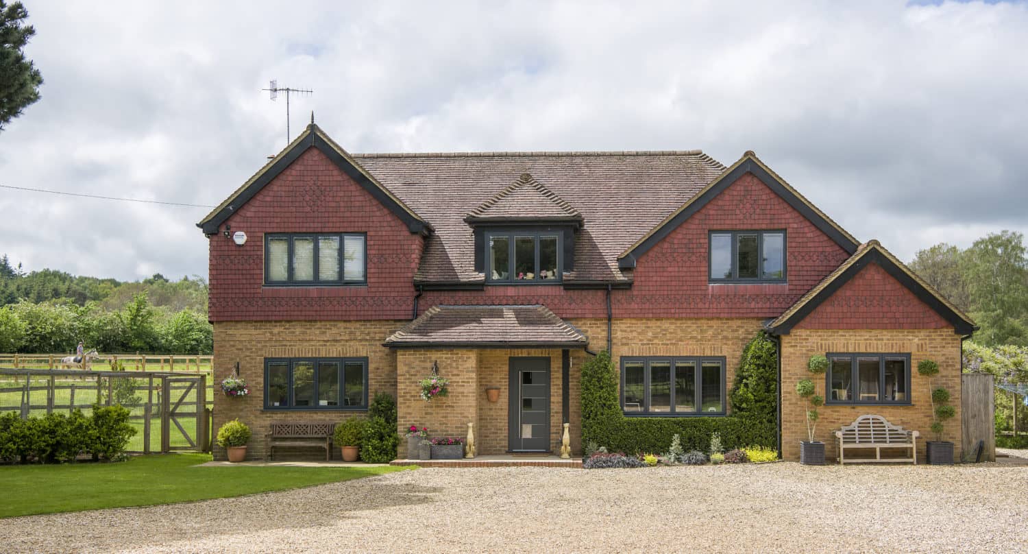 Two-story house with red and yellow brick exterior, twin gables, grey front door, multiple windows, gravel driveway, green lawn, benches, flowerpots, and wooden gate, set against a backdrop of trees and cloudy sky. Features glass roofing over the entryway.