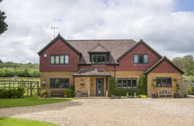 Two-story house with red and yellow brick exterior, twin gables, grey front door, multiple windows, gravel driveway, green lawn, benches, flowerpots, and wooden gate, set against a backdrop of trees and cloudy sky. Features glass roofing over the entryway.