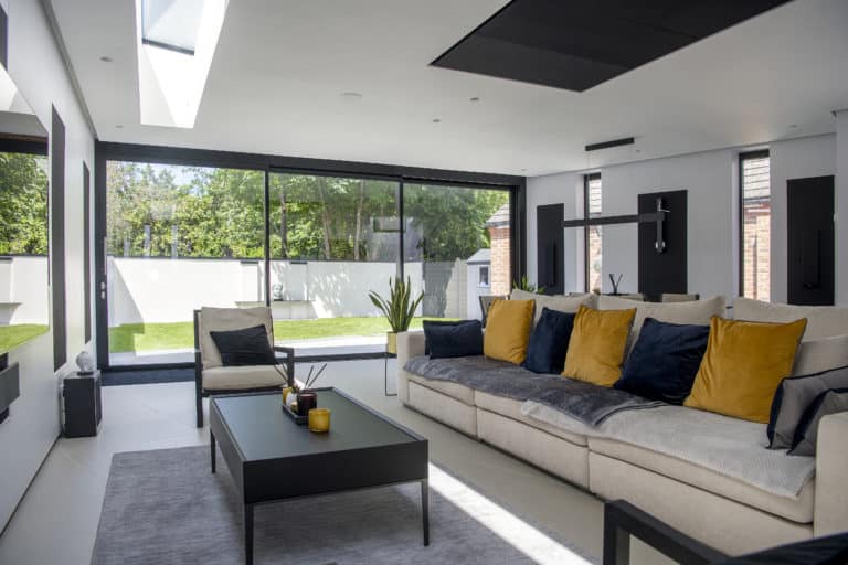 A modern living room with a beige sectional sofa decorated with blue and yellow cushions, a black coffee table, large floor-to-ceiling windows, and bespoke glass solutions offering a view of the grassy backyard and trees outside.