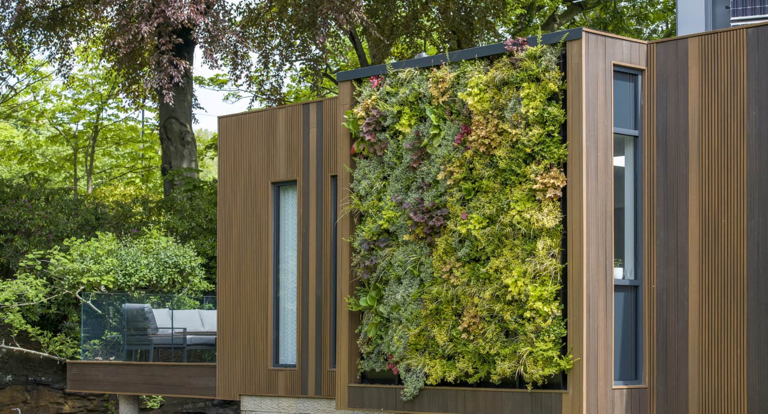 Modern house exterior with a large vertical green wall filled with various plants and foliage, surrounded by wood panels; sleek bifold doors open to a patio area with outdoor seating, seamlessly connecting indoor and outdoor spaces among trees.