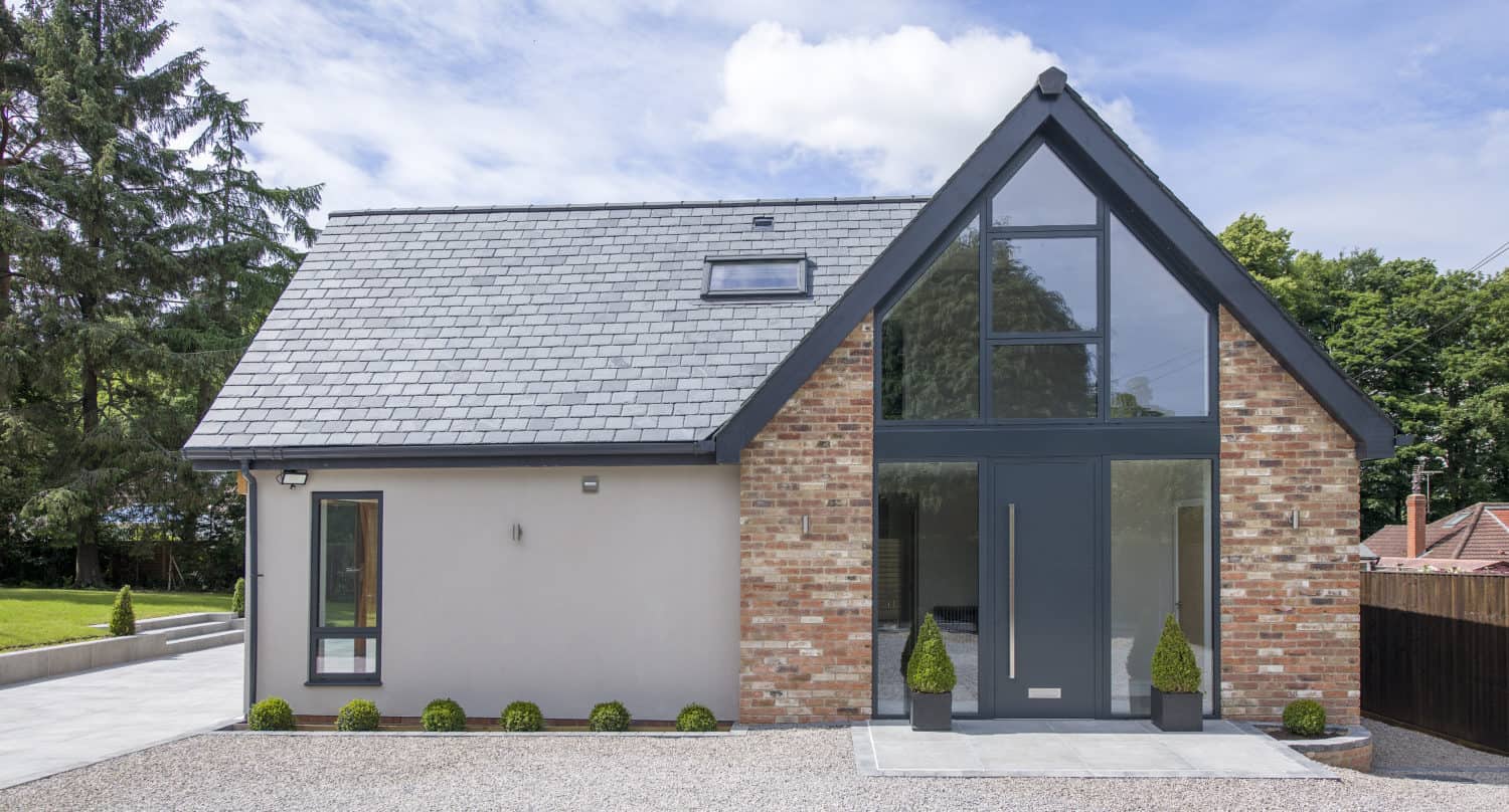 Modern house with a pitched slate roof, brick and light-colored walls, large floor-to-ceiling windows and stylish bifold doors at the front, plus neat landscaping with small shrubs on a gravel driveway.