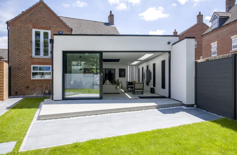 A modern house extension with large sliding doors, white walls, a flat roof, and minimalist decor opens onto a patio and well-kept lawn, surrounded by traditional brick houses. Glass roofing adds brightness to the sleek design.