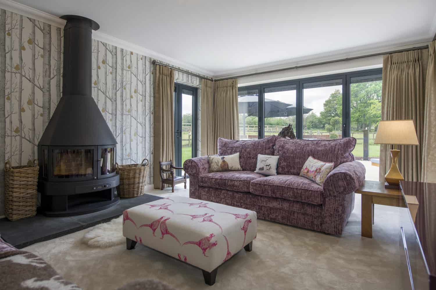 A cozy living room with a purple sofa, floral ottoman, modern black wood-burning stove, patterned wallpaper, large windows, and bifold doors opening to views of a green lawn and trees outside.
