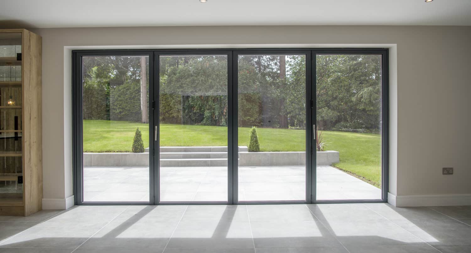 Large floor-to-ceiling bifold doors open to a patio and a green garden with trees and manicured grass, letting natural light flood the modern, tiled interior space.
