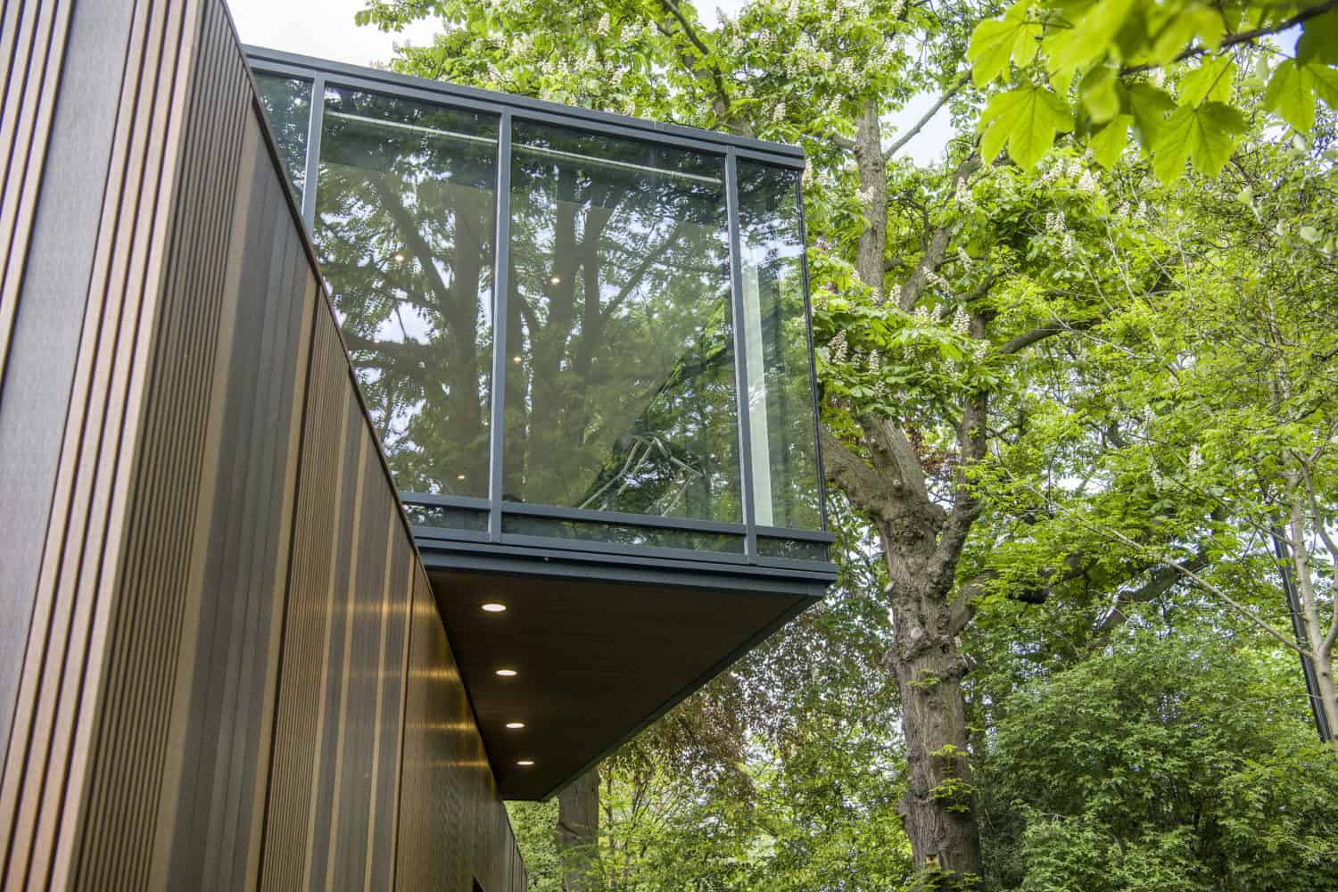Modern building with a cantilevered glass room and bifold doors, surrounded by lush green trees reflecting in the glass. The lower wood-paneled wall features recessed lights. The scene appears bright and natural.