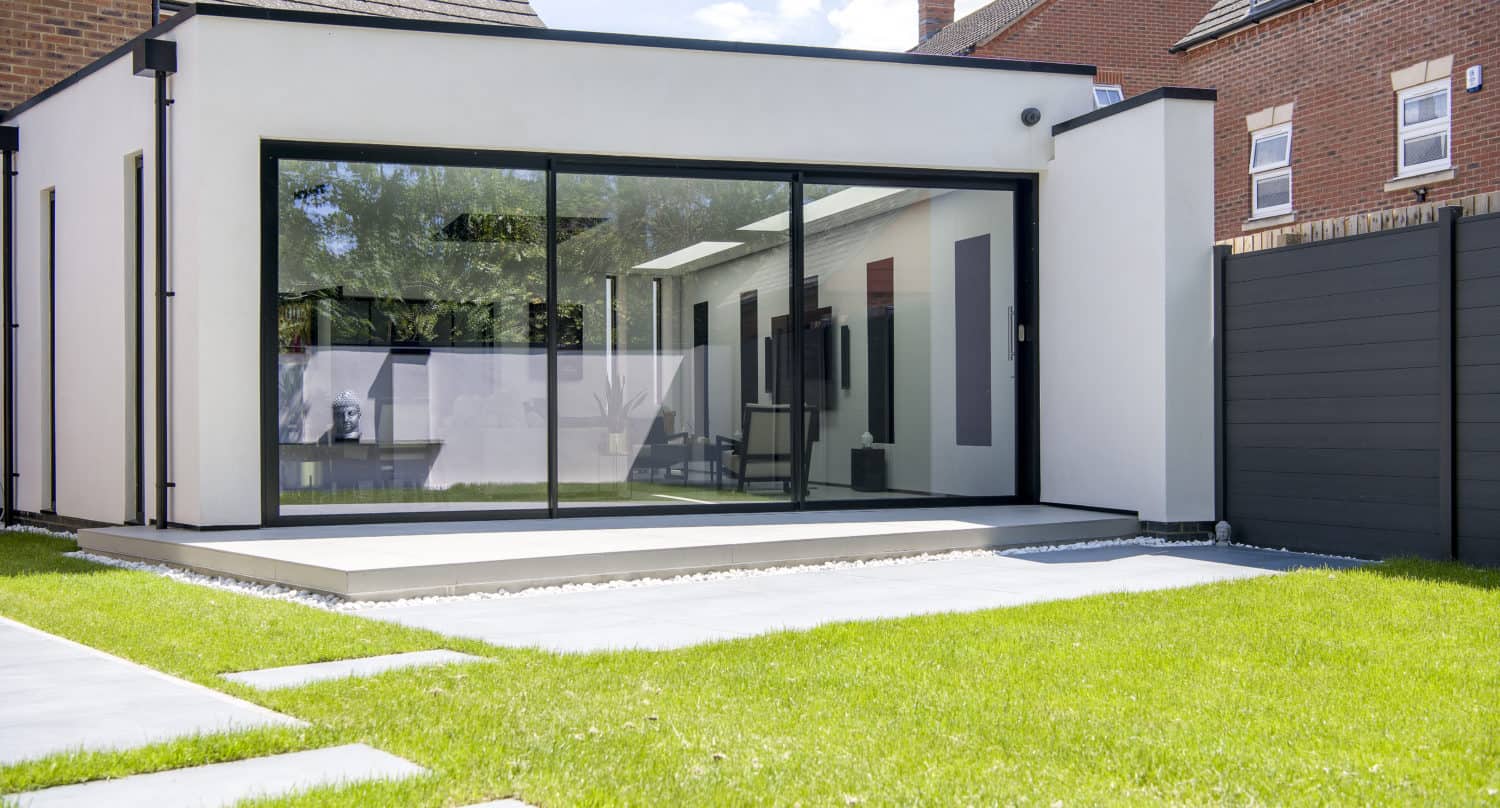 Modern home extension with large glass sliding doors, white walls, and a flat roof. The building opens onto a patio and green lawn, with neighboring brick houses visible in the background. Stylish bifold doors enhance the seamless indoor-outdoor flow.