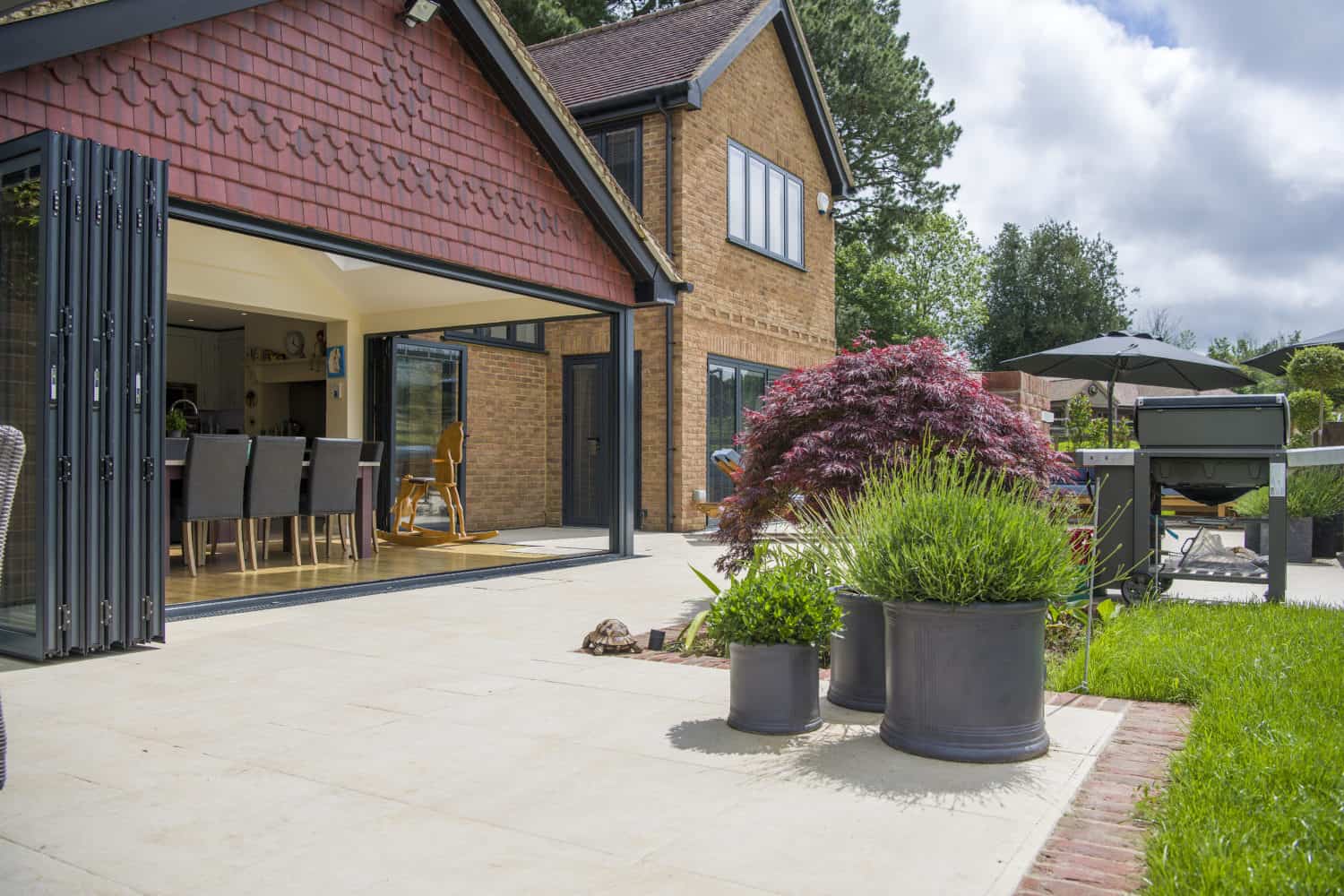 A modern house with bespoke glass solutions, including large glass folding doors opening to a sunny patio with potted plants, outdoor dining furniture, and a garden featuring green grass and trees in the background.
