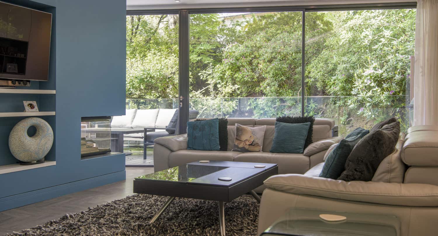 Modern living room with gray sofas, teal accents, a glass coffee table on a shaggy rug, large windows and sliding doors showcasing lush greenery outside, and a blue wall with built-in shelves and a TV.