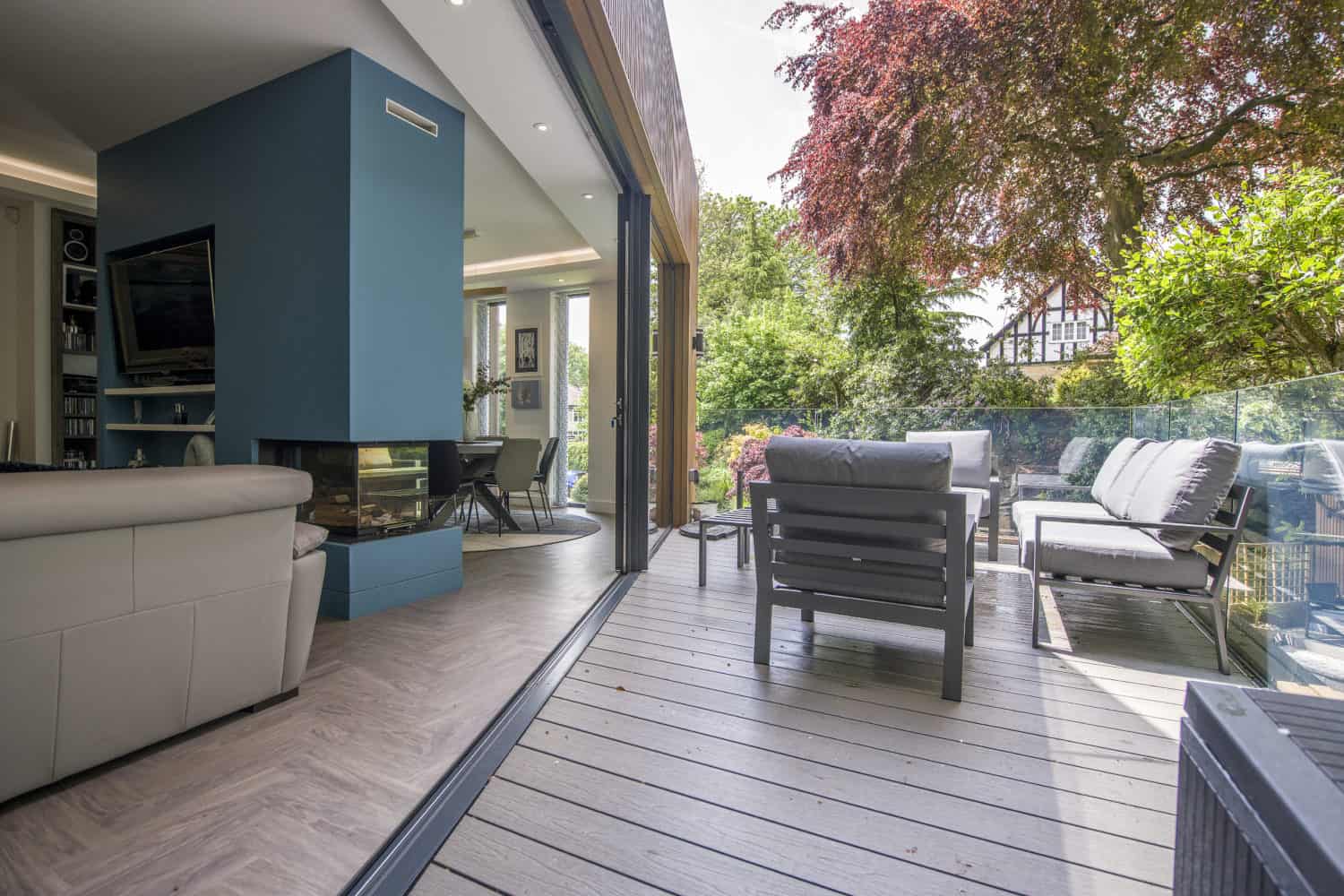 A modern living room with light-colored furniture opens onto a spacious patio with seating. Bifold doors connect indoor and outdoor areas, overlooking a lush garden filled with trees and greenery.