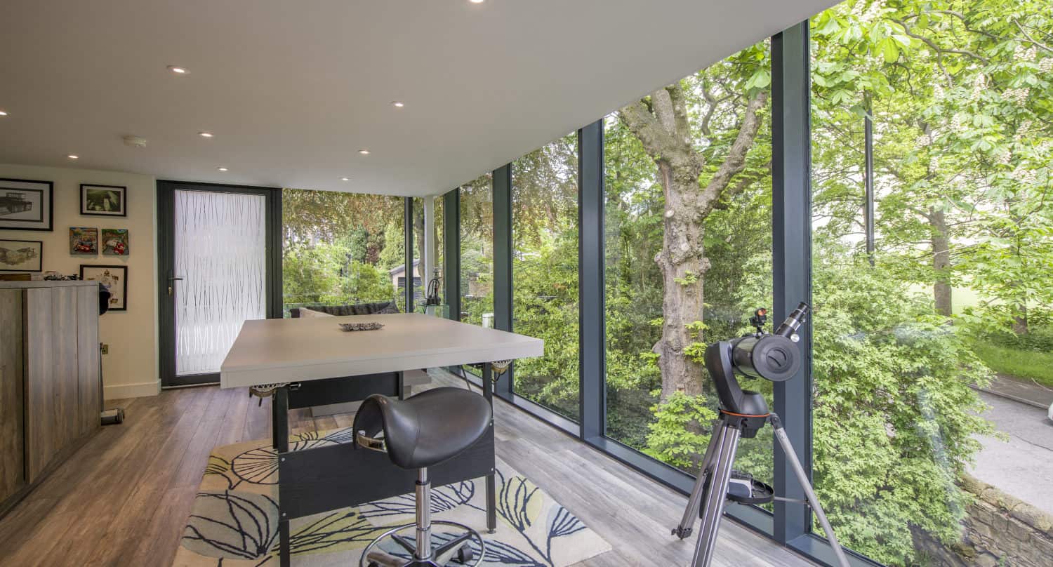 Modern room with floor-to-ceiling windows and bespoke glass solutions overlooking lush trees, featuring a white desk, black chair, and telescope. Bright natural light fills the space with wooden flooring and a patterned rug.