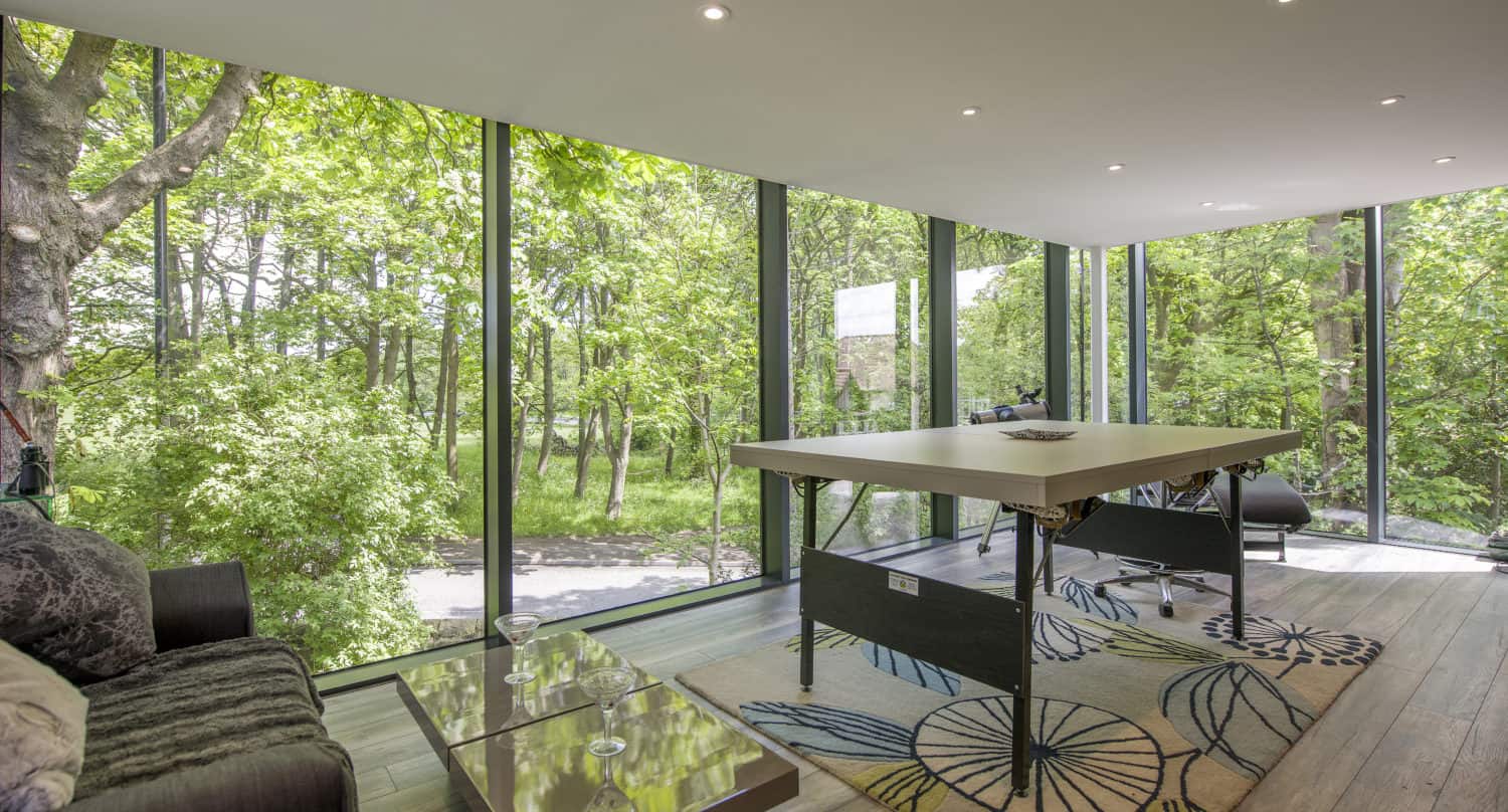 A modern room with floor-to-ceiling bespoke glass solutions overlooks a lush green forest. Inside, there is a ping pong table on a patterned rug, a black couch, and a glass coffee table on wood flooring.