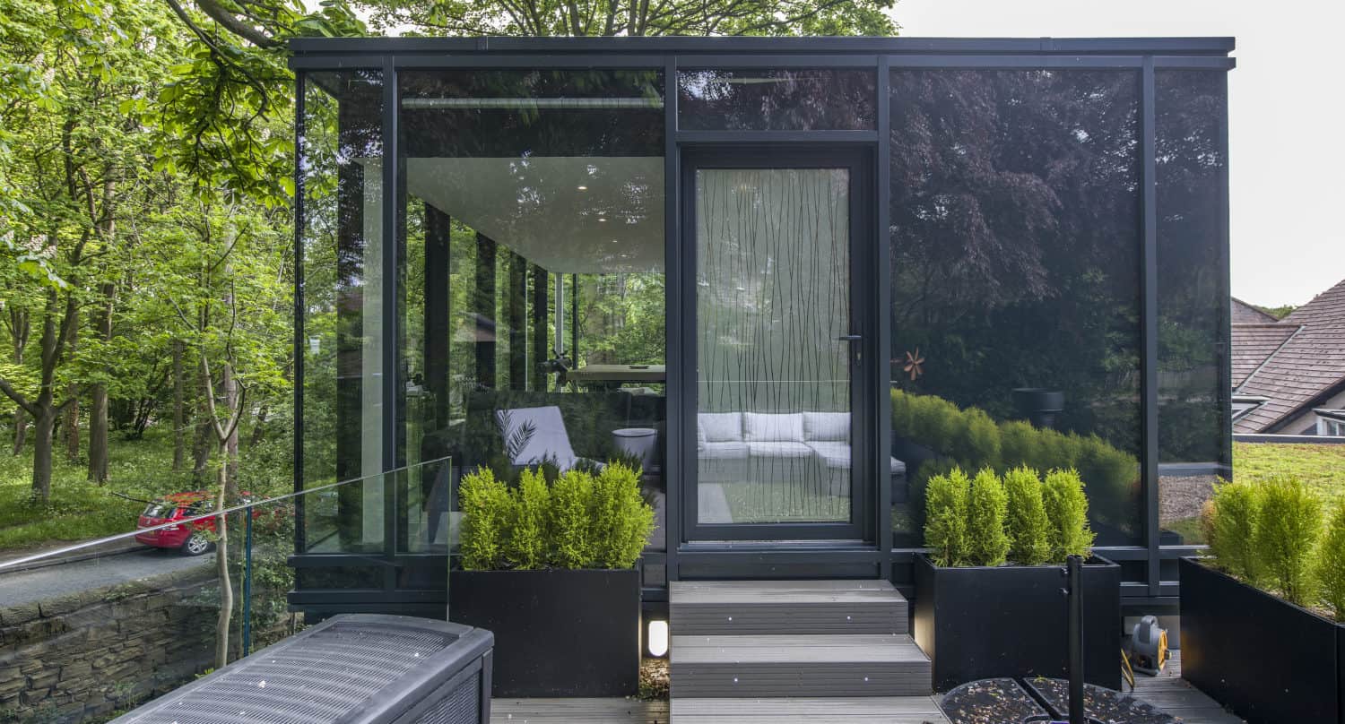 Modern glass-walled garden room with bespoke glass solutions and black trim, surrounded by greenery. Steps lead up to the door, and tall plants line the entrance. Trees and a road with a red car are visible in the background.