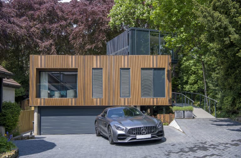 A modern house with a wood-paneled exterior and large windows, featuring sliding doors, sits among trees. A gray luxury sports car is parked in front of its closed garage on a paved driveway.