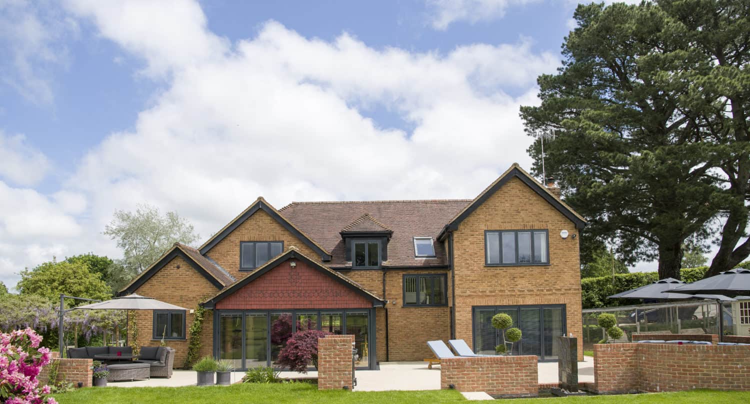 A modern two-story brick house with large windows, sliding doors, a landscaped garden, outdoor seating areas, and trees under a partly cloudy sky.