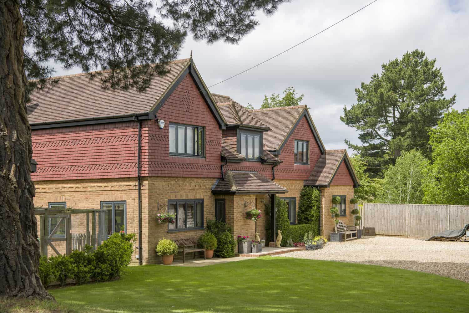 A large brick and red-tiled house with multiple gables, black-framed windows, bifold doors, potted plants, hanging flower baskets, and a well-kept lawn, surrounded by trees and a gravel driveway under a partly cloudy sky.