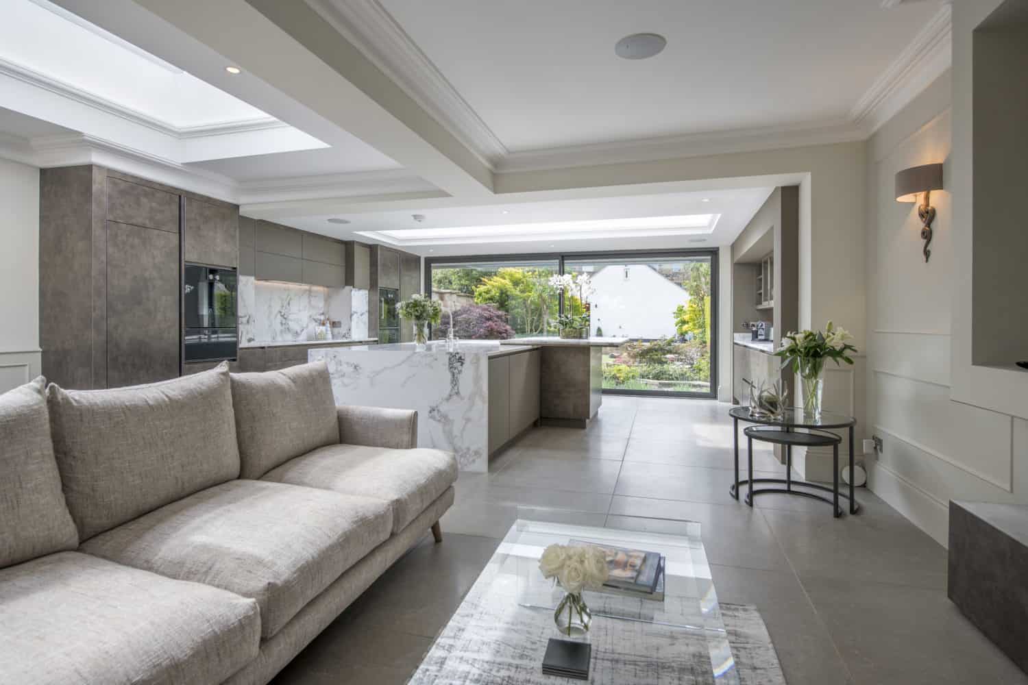 Modern open-plan living space with a light beige sofa, glass coffee table, and a stylish kitchen featuring marble countertops. Large bespoke glass solutions, including sliding front doors, open to a lush garden and flood the area with natural light.