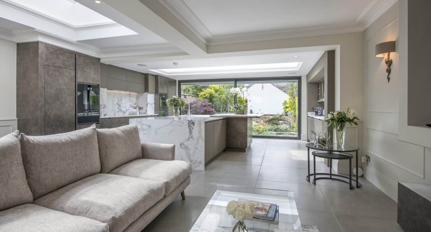Modern open-plan living space with a light beige sofa, glass coffee table, and a stylish kitchen featuring marble countertops. Large bespoke glass solutions, including sliding front doors, open to a lush garden and flood the area with natural light.