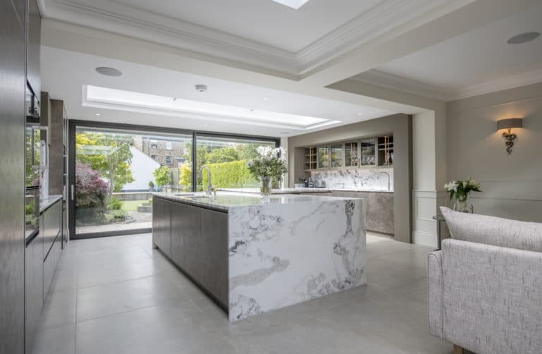 Modern kitchen with a large marble island, sleek cabinets, and floor-to-ceiling sliding doors opening to a lush green garden. Natural light fills the space, creating a bright and airy atmosphere.