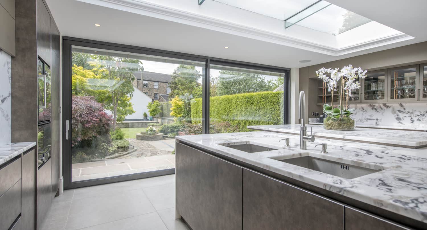 Modern kitchen with marble countertops and two sinks, large skylight, and glass sliding doors opening to a lush green garden with trees and bushes. Bright natural light fills the space through the sliding doors.