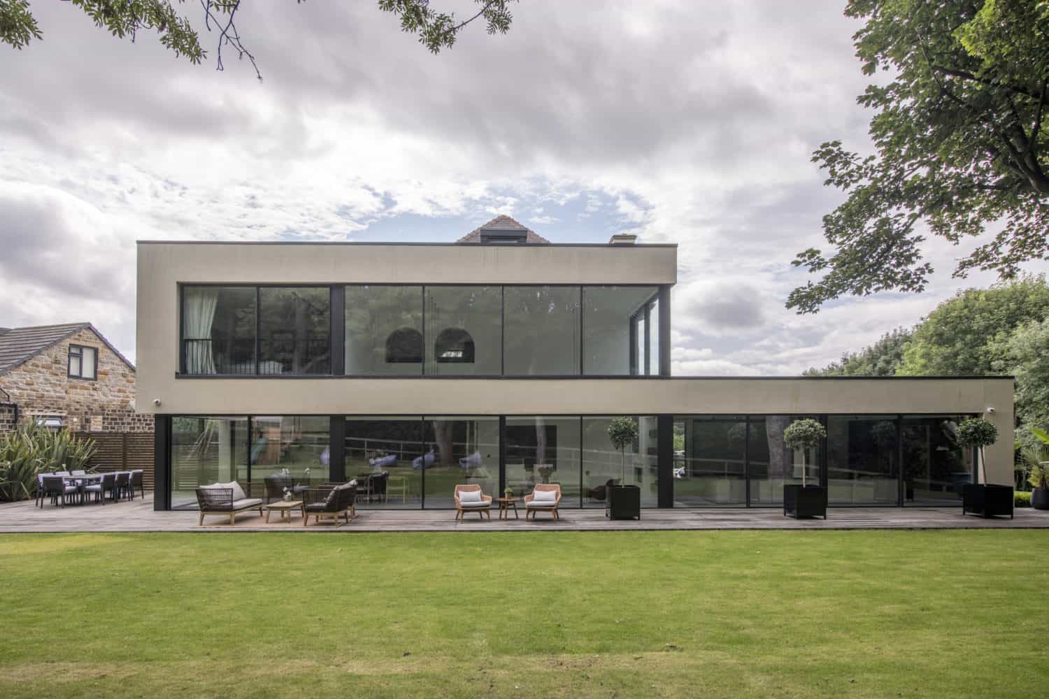 Modern two-story house with bespoke glass solutions, featuring large windows, a spacious wooden patio with outdoor seating, and a well-kept lawn in the foreground. Trees and greenery surround the contemporary building.