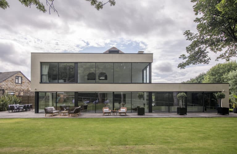Modern two-story house with bespoke glass solutions, featuring large windows, a spacious wooden patio with outdoor seating, and a well-kept lawn in the foreground. Trees and greenery surround the contemporary building.