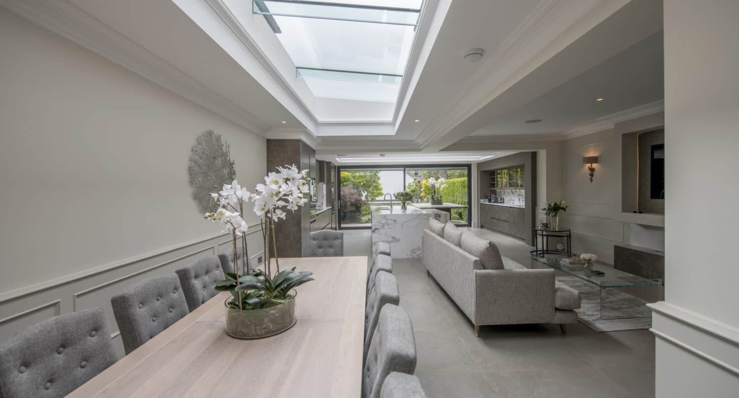 Modern living and dining area with neutral-toned furniture, large glass roofing, bifold doors opening to a garden, and potted orchids on the dining table, creating a bright, airy atmosphere.