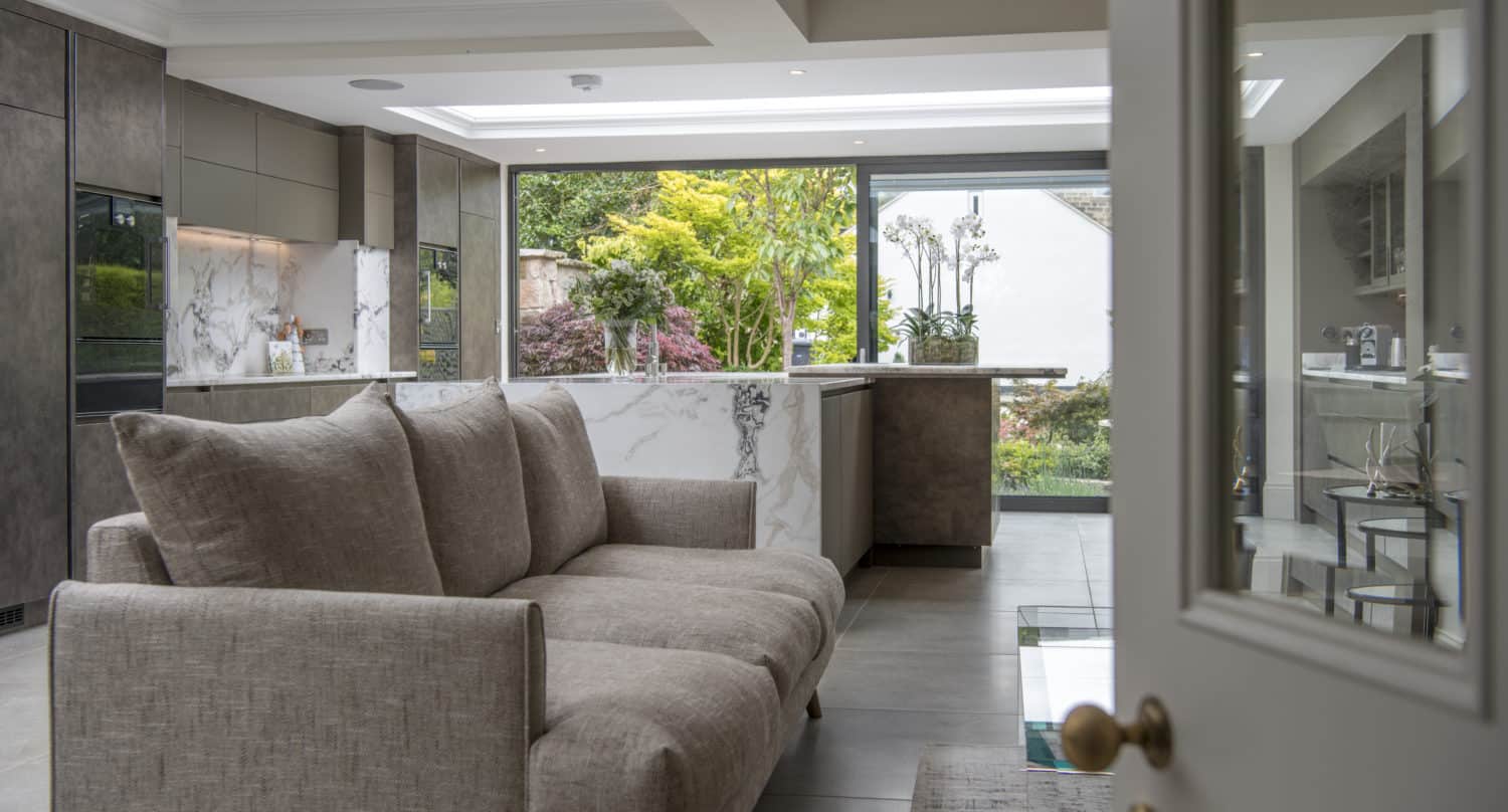 A modern open-plan kitchen and living area with a beige sofa, marble countertops, and large windows featuring bespoke glass solutions. Natural light fills the space, highlighting contemporary decor and lush garden views.
