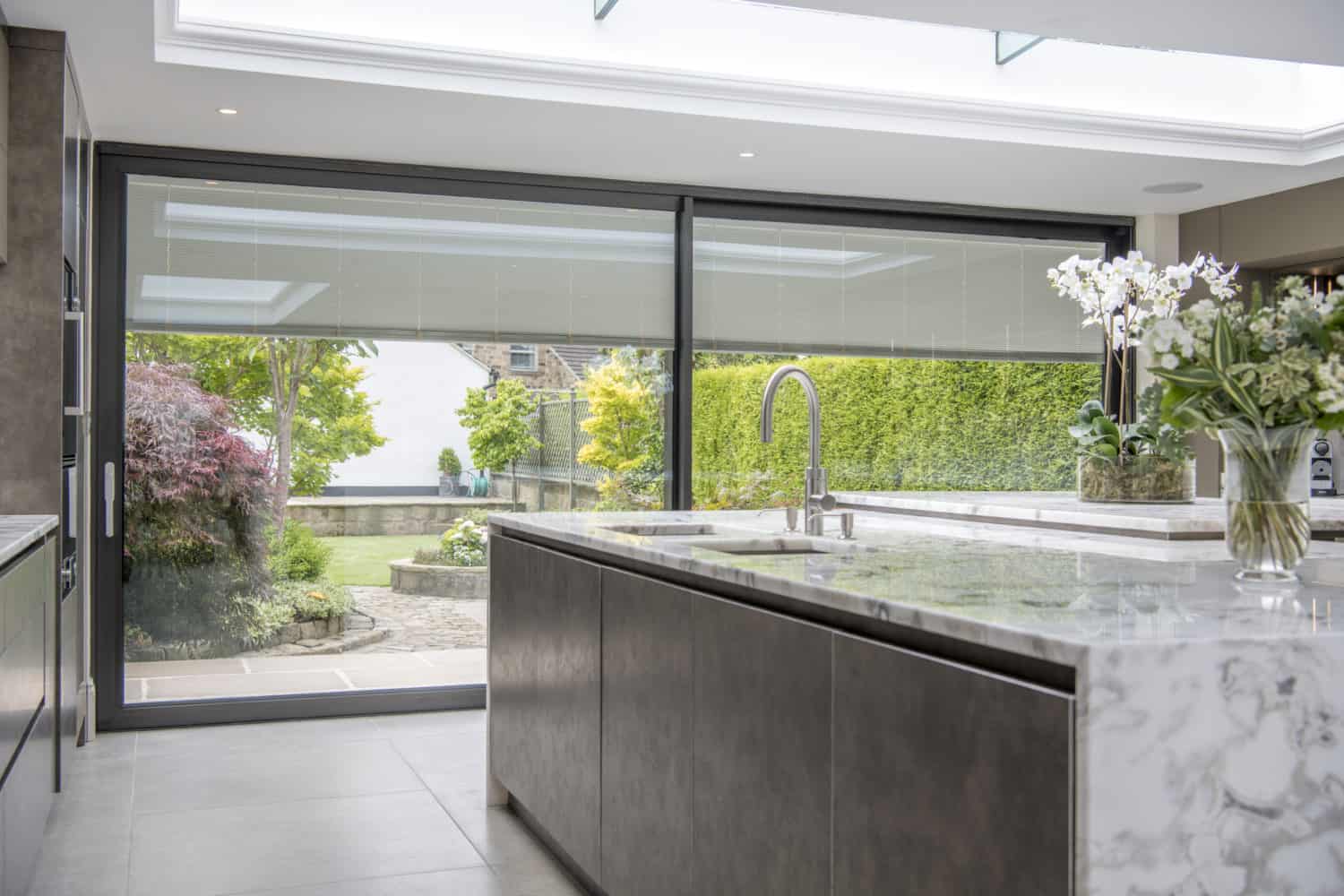 Modern kitchen with a marble island, built-in sink, and bespoke sliding doors leading to a landscaped garden. Natural light streams in through a skylight, highlighting potted orchids on the countertop.